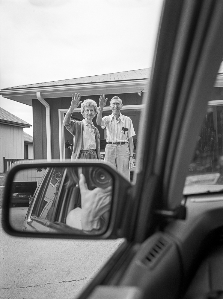 Black and white photograph taken from inside a car, showing an elderly woman and man waving from the porch of a mid-century suburban house. The composition frames the subjects through the car's side mirror and open window, creating layered depth between the foreground and the figures in the distance.