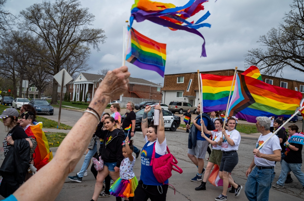 A crowd of people participates in a Pride parade on a residential street, holding and waving rainbow flags of various designs. Participants in colorful clothing move through the scene as bare trees and suburban houses frame the background under an overcast sky.