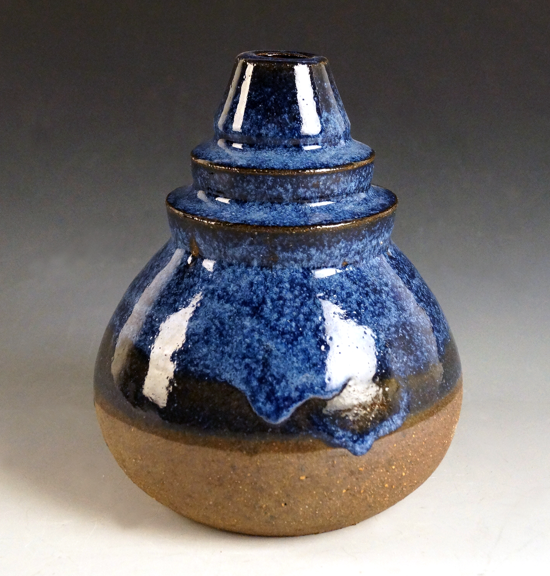 A ceramic vessel with a bulbous body and tapered neck features layered glazing in deep blue, white, and warm brown tones. The textured surface shows dripping glaze effects and metallic accents at the rim, creating an organic, gestural quality against the earthenware base.