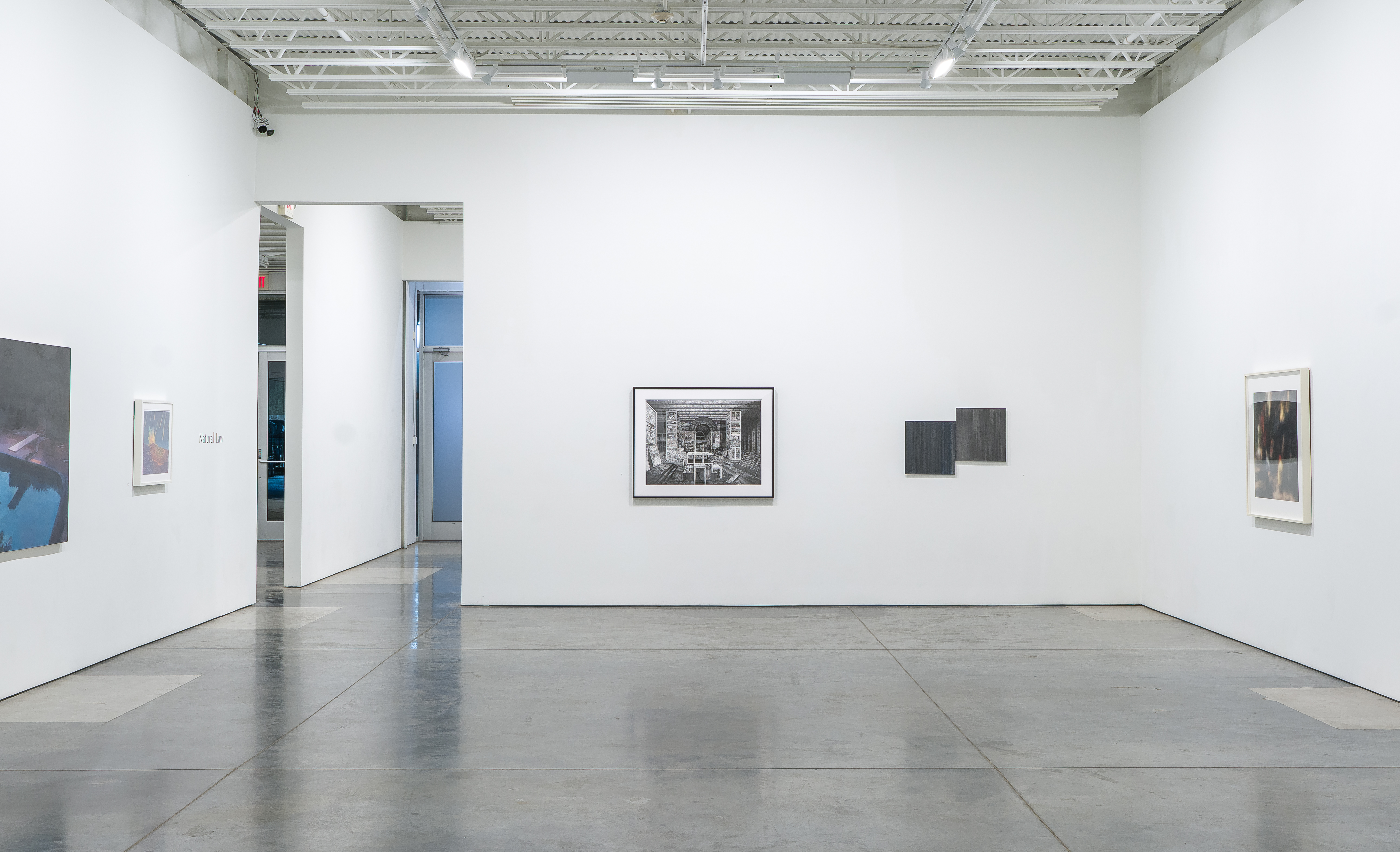 A minimalist gallery interior with white walls and polished concrete flooring. Several framed artworks are sparsely distributed across the walls at varying heights, including a detailed architectural print, dark abstract pieces, and photographic works in white frames. The space features industrial ceiling elements and natural light, emphasizing the scale and openness of the gallery.