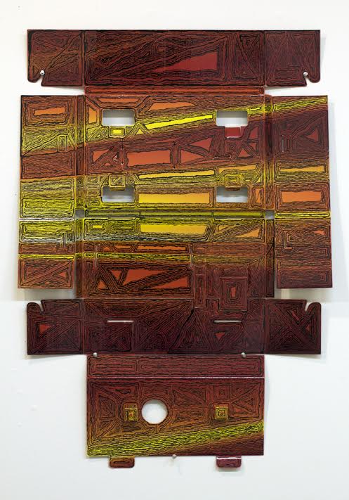 Abstract geometric composition made from layered cardboard pieces in warm earth tones—deep reds, browns, and yellows—with incised linear patterns creating a grid-like structure. White rectangular cutouts and openings punctuate the central composition, arranged in an asymmetrical, roughly symmetrical overall form against a white wall.