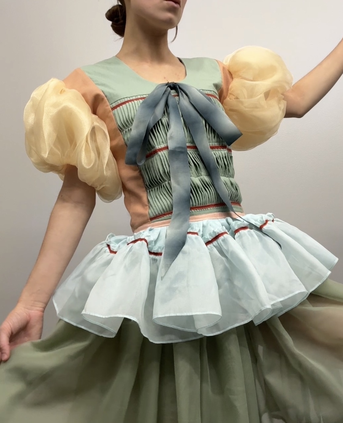 A figure wears a structured ensemble with a sage green pleated bodice accented by a gray ribbon bow, paired with a layered skirt featuring white organza ruffles and a sage green underskirt trimmed in red. Voluminous cream-colored puffed sleeves frame the composition against a neutral background.