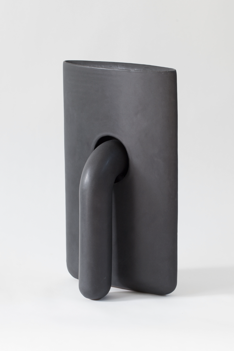 Abstract ceramic sculpture in dark gray stoneware featuring a rectangular vertical form with a curved tubular element protruding from its front, creating an interplay of positive and negative space against a neutral background.