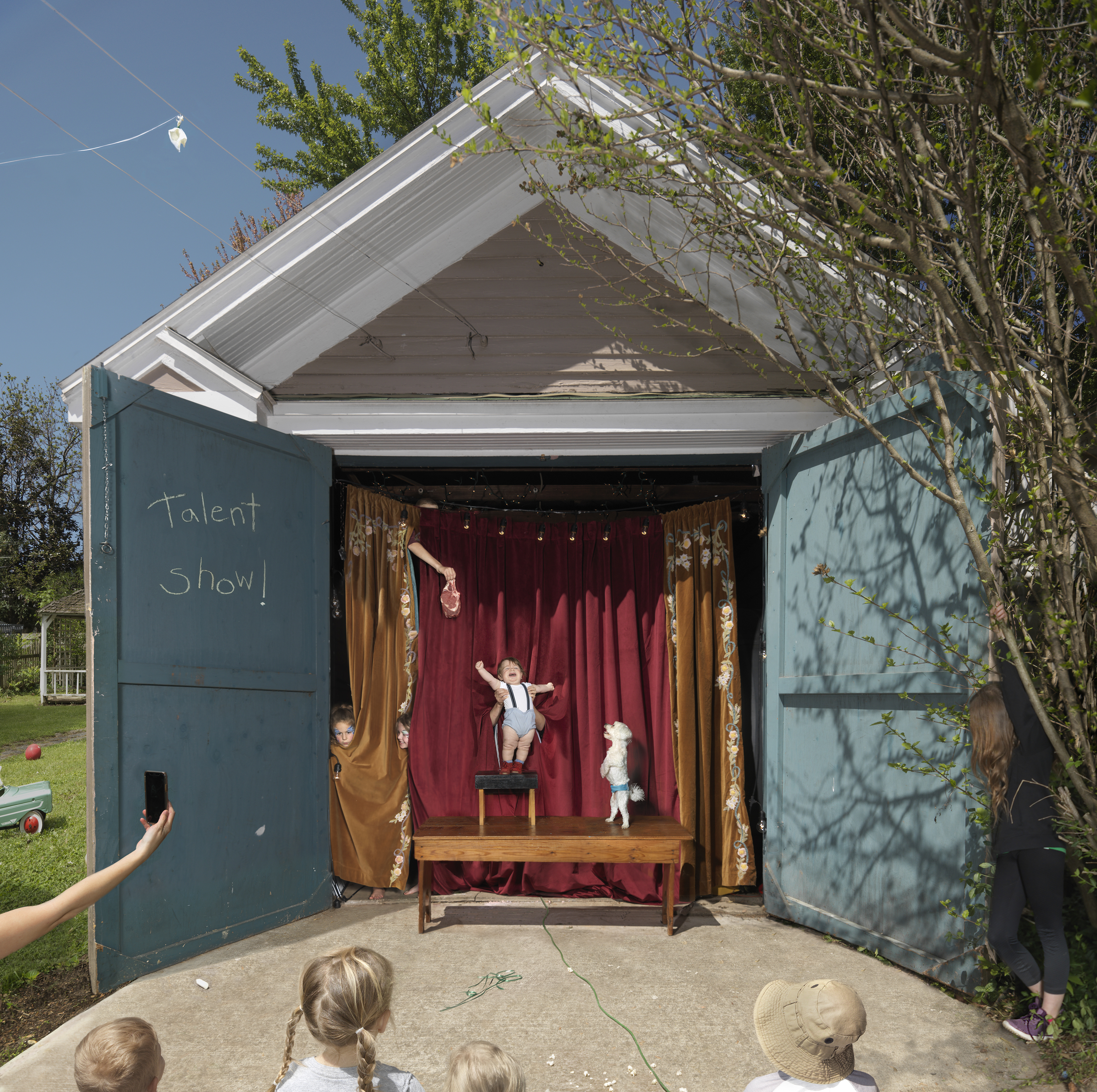 A makeshift outdoor theater constructed from a garden shed features open doors revealing a stage with burgundy curtains, gold drapes, and a wooden platform. Children perform acrobatic acts on the stage while an audience of young viewers watches from outside, with one child documenting the scene on a phone. The word "Talent Show" is written in chalk on the shed's exterior wall.