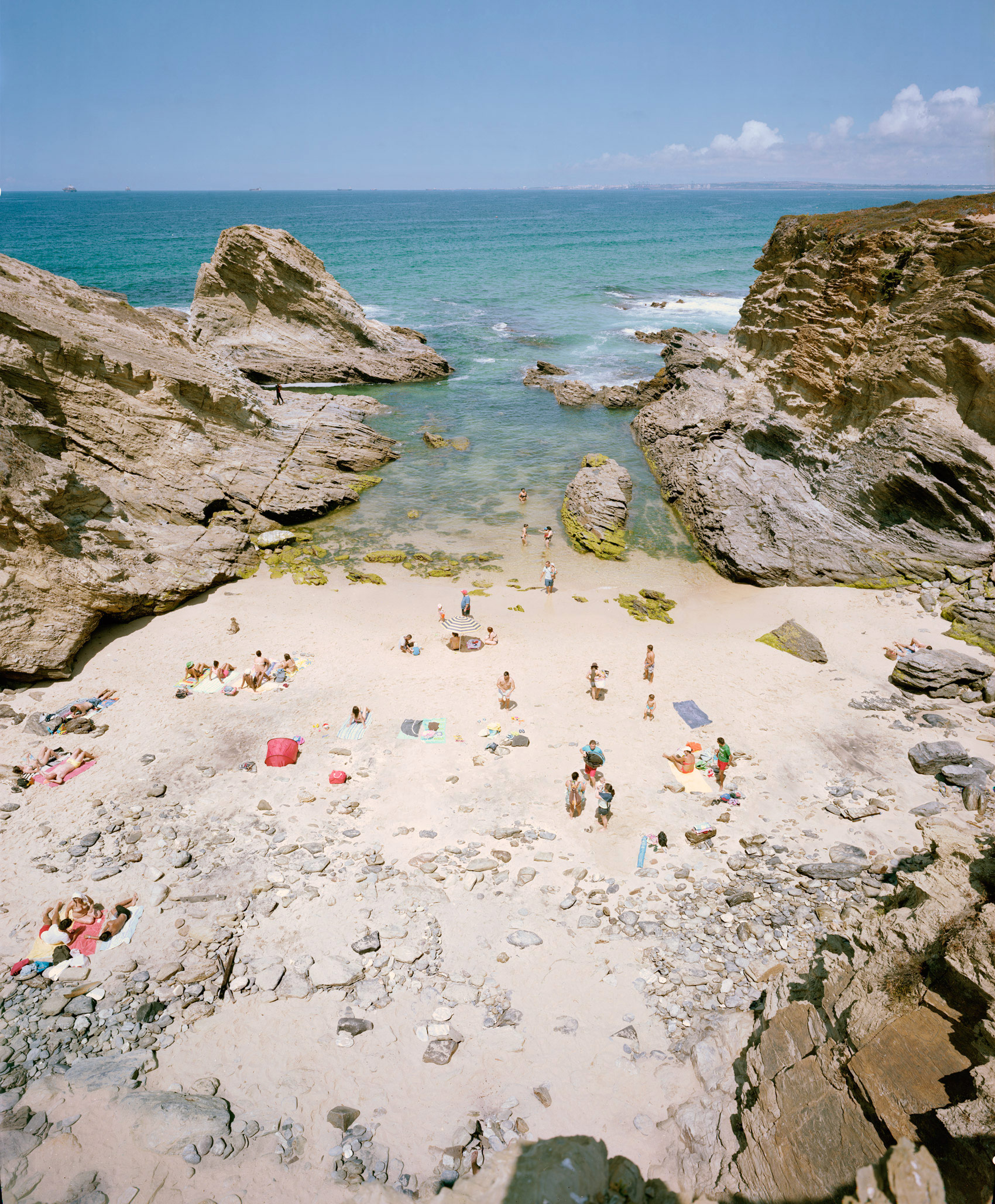 Aerial view of a sheltered sandy cove framed by dramatic stratified limestone cliffs, with turquoise water and scattered beachgoers in summer attire. Moss-covered rocks dot the pale sand, while the horizon shows calm seas under a partly cloudy sky.