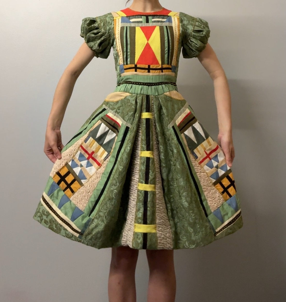 A dress constructed from patchworked upholstery and cotton fabric in sage green, featuring bold geometric appliqués in red, yellow, black, and cream across the bodice and full skirt. Puffed sleeves and cutout details at the waist emphasize the garment's sculptural construction, while intricate embroidered patterns and linear designs create a striking visual rhythm throughout.