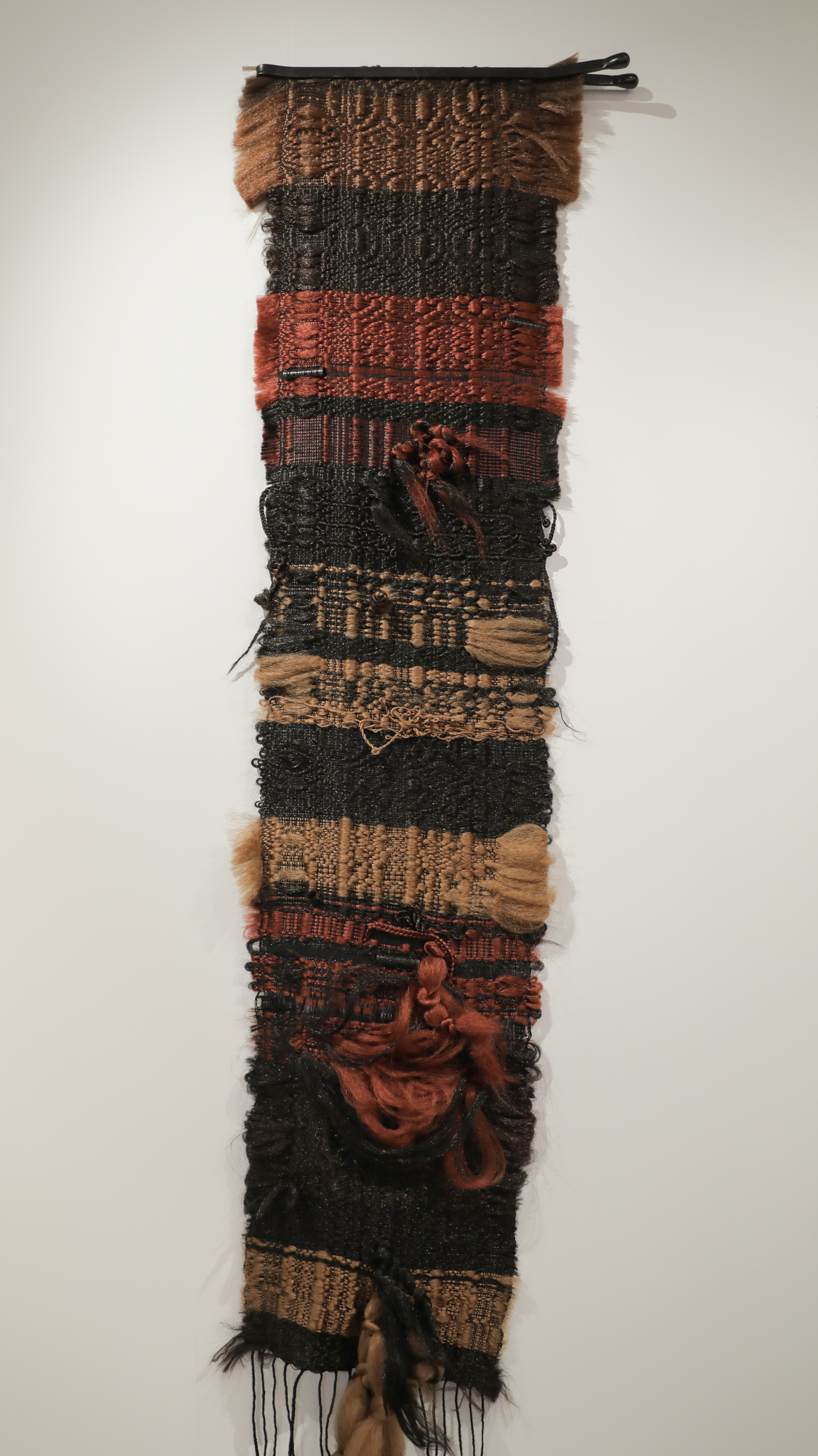 A vertical textile artwork composed of stacked fabric strips in varying weaves and textures, featuring earthy tones of brown, black, rust red, and tan. The composition includes woven sections, synthetic hair elements, and sculptural components including plastic rollers and bobby pins integrated throughout, with loose fringe at the bottom.
