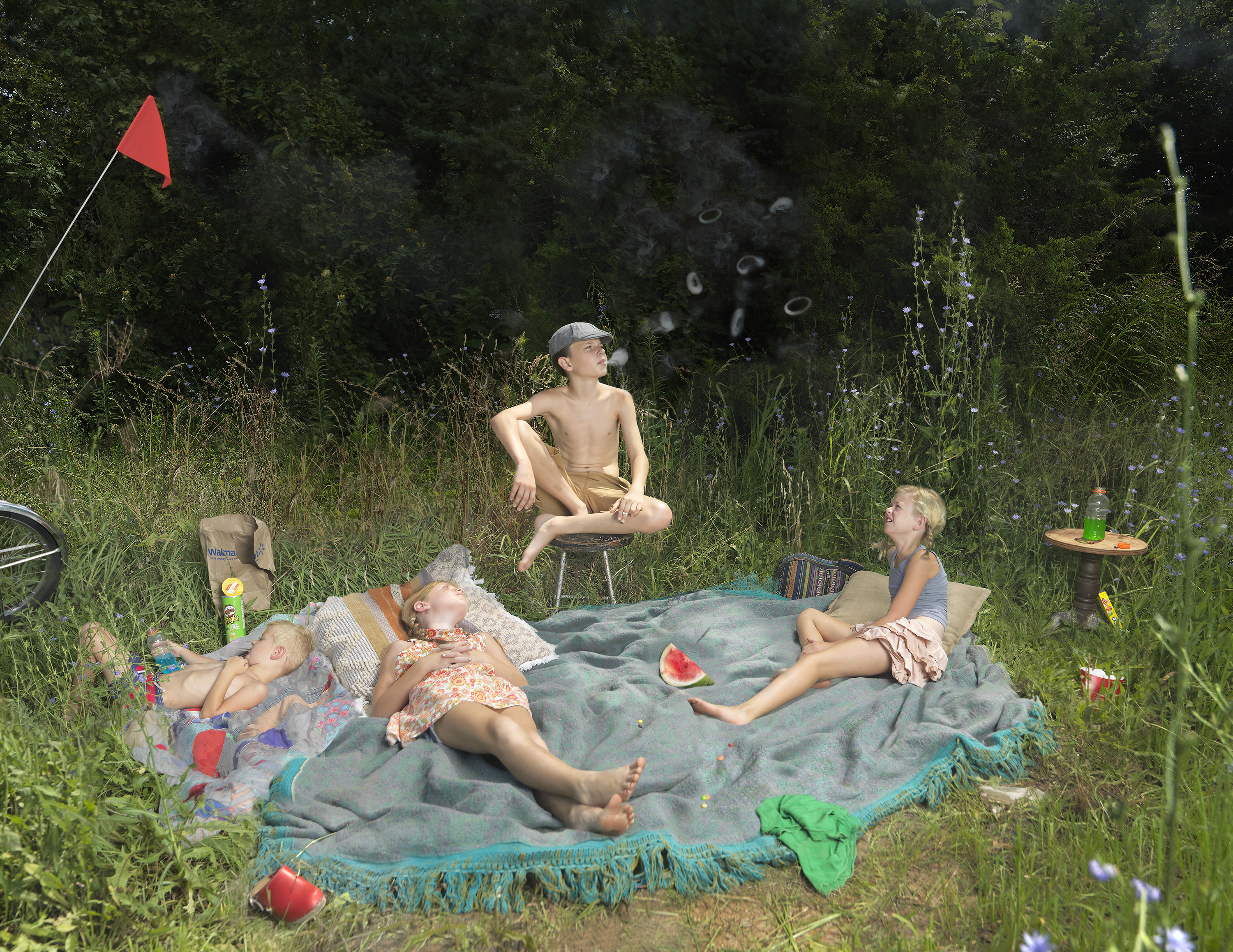 A family picnic scene set in an overgrown field surrounded by tall grass and wildflowers. Four children of varying ages relax on a blue blanket spread across the ground, with a watermelon slice visible among snacks and supplies. A red flag marks the picnic spot in the upper left, while a shirtless boy crouches on a camp chair observing the scene. The composition blends domestic leisure with natural wilderness, rendered in saturated color and documentary-style photography.