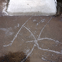 White chalk lines drawn on a wet concrete surface, creating abstract curved and intersecting marks. A white rectangular object, possibly a cushion or pad, is visible at the top edge of the frame.
