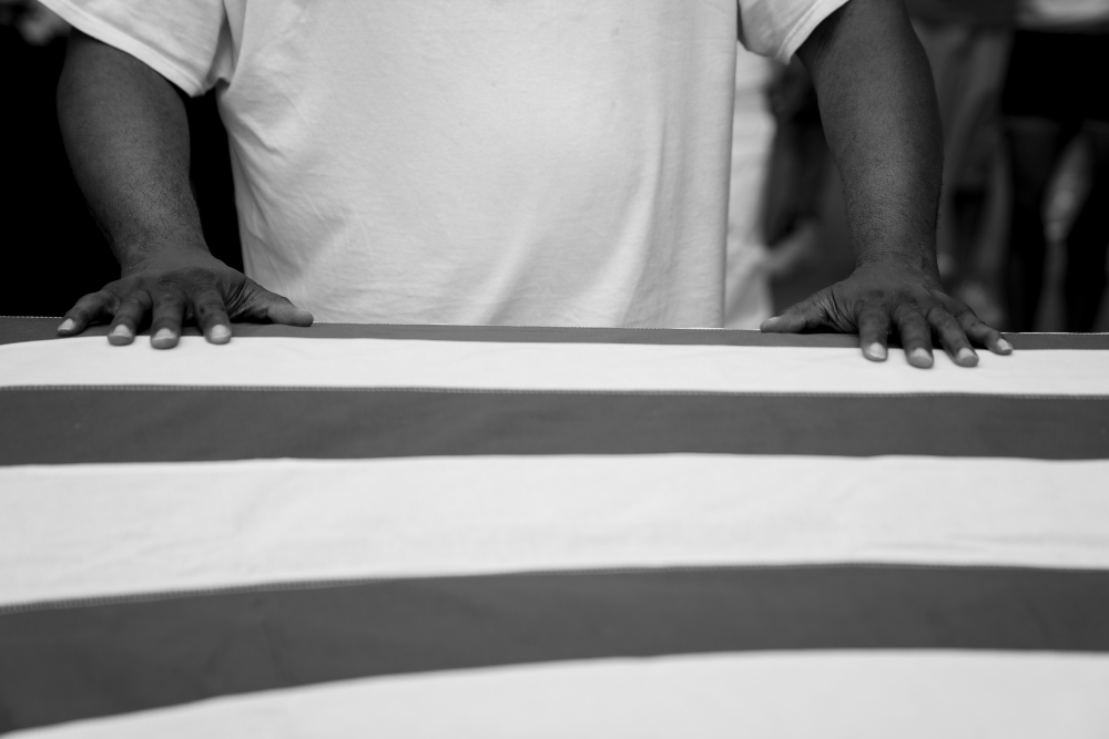 Black and white photograph of a person wearing a white t-shirt with both hands placed on a curved white surface with dark horizontal stripes, captured from a frontal perspective emphasizing gesture and compositional geometry.