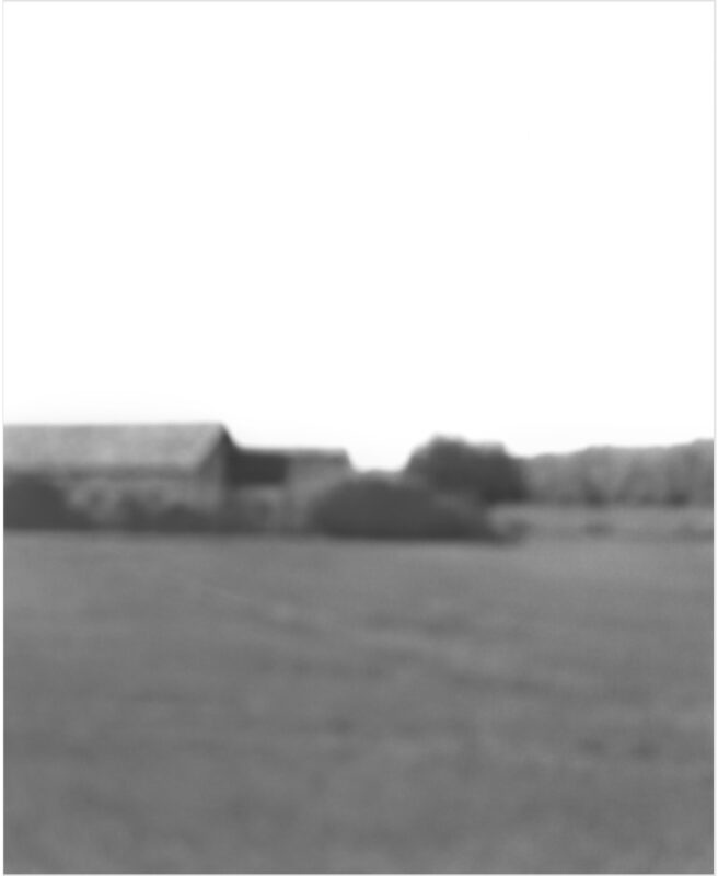 Monochromatic landscape photograph featuring a distant rural scene with farm buildings and trees along the horizon, rendered in soft focus with a hazy, atmospheric quality. The composition is dominated by an empty foreground of flat terrain beneath a pale sky.