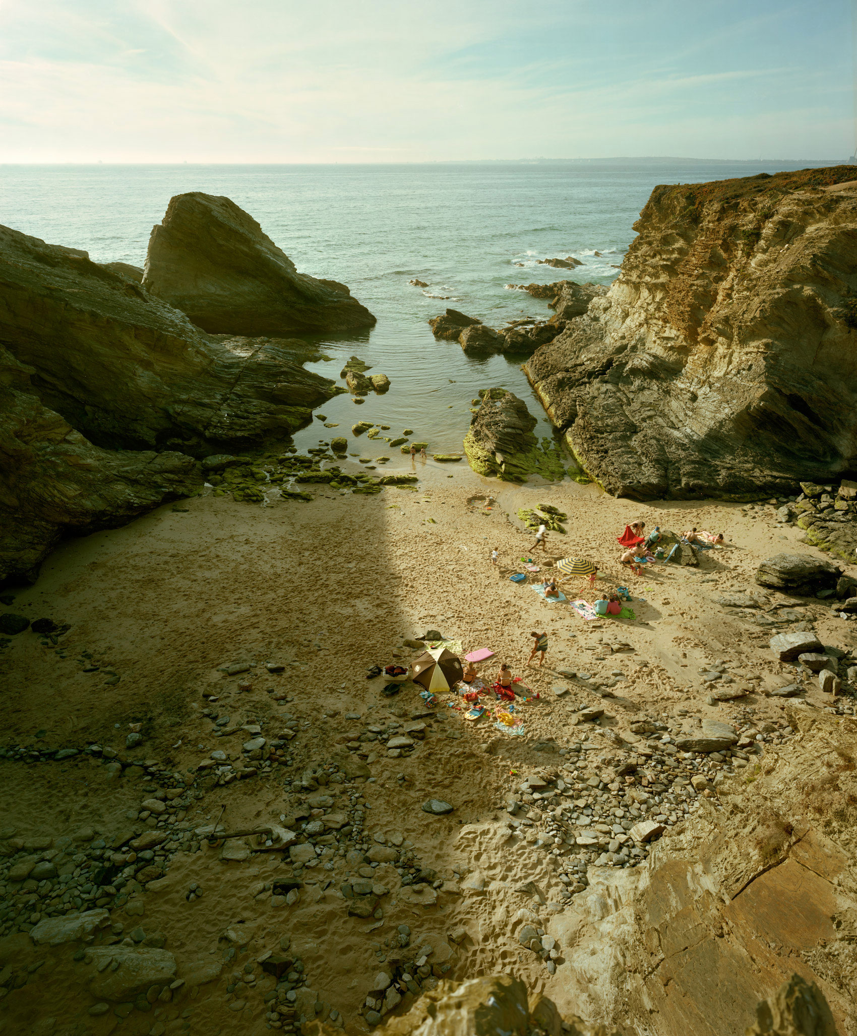 A secluded rocky cove with moss-covered cliff formations framing a sandy beach where beachgoers with colorful umbrellas and towels are scattered across the shore, turquoise water visible beyond.