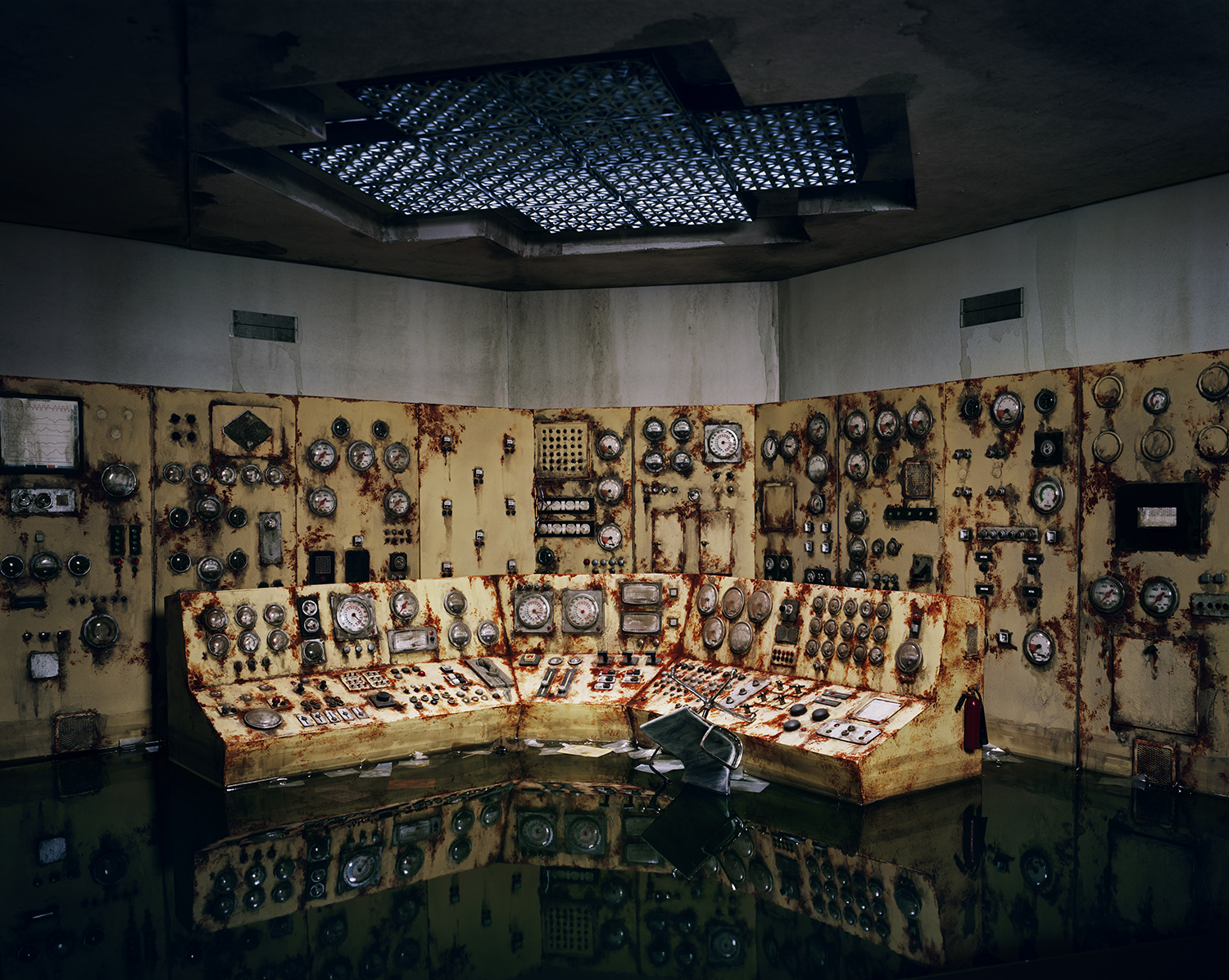A deteriorated control room interior with rust-stained yellow panels densely covered in gauges, dials, switches, and monitoring equipment. Corroded console desks arranged in an L-shape occupy the center, while matching instrumentation panels line the walls and ceiling, all reflecting in a flooded floor below.