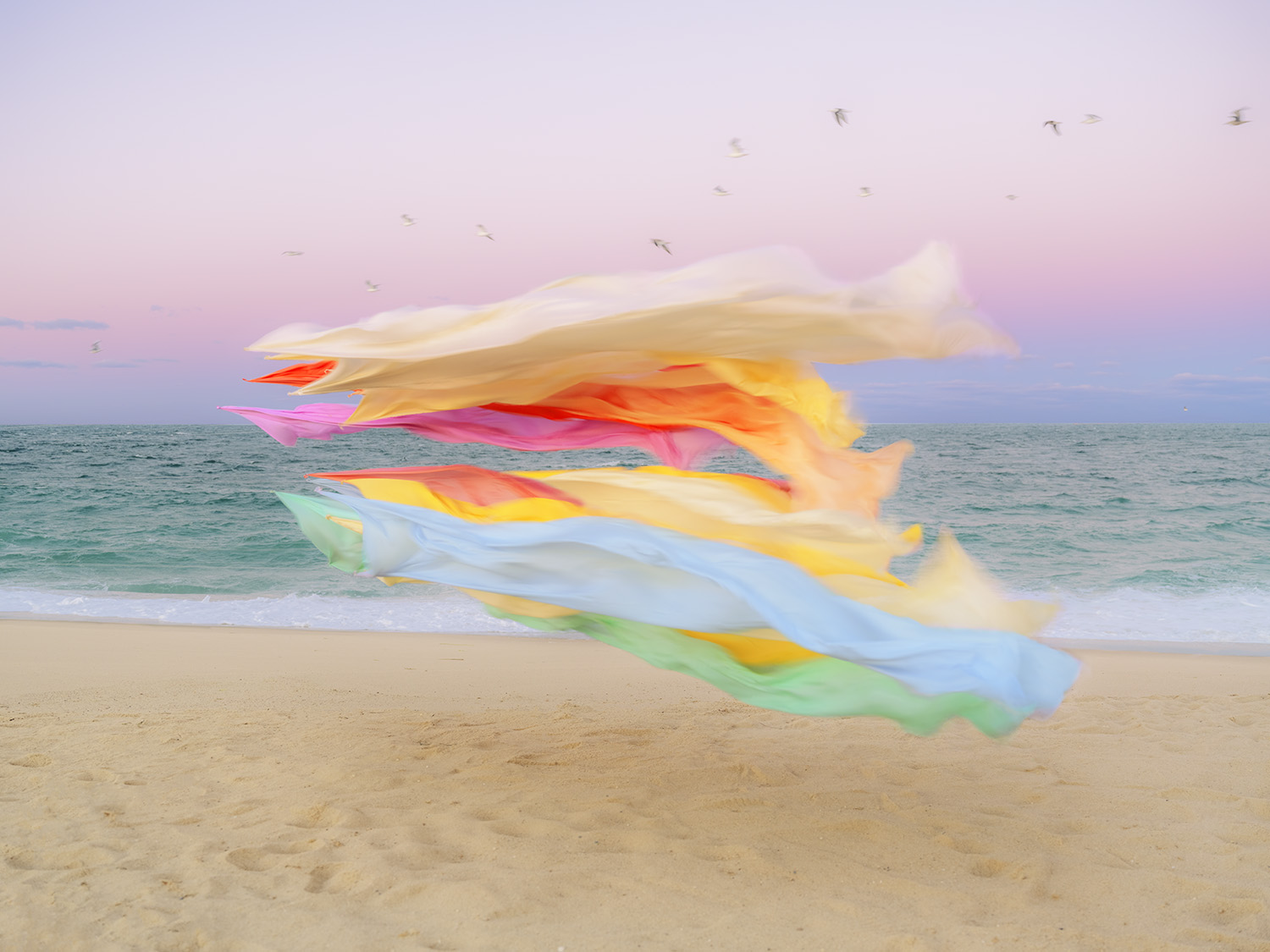 Billowing sheets of colorful silk—layered in shades of white, cream, pink, red, orange, yellow, blue, and green—dance in the wind on a sandy beach at dusk, with the ocean and a purple-pink sky visible beyond.