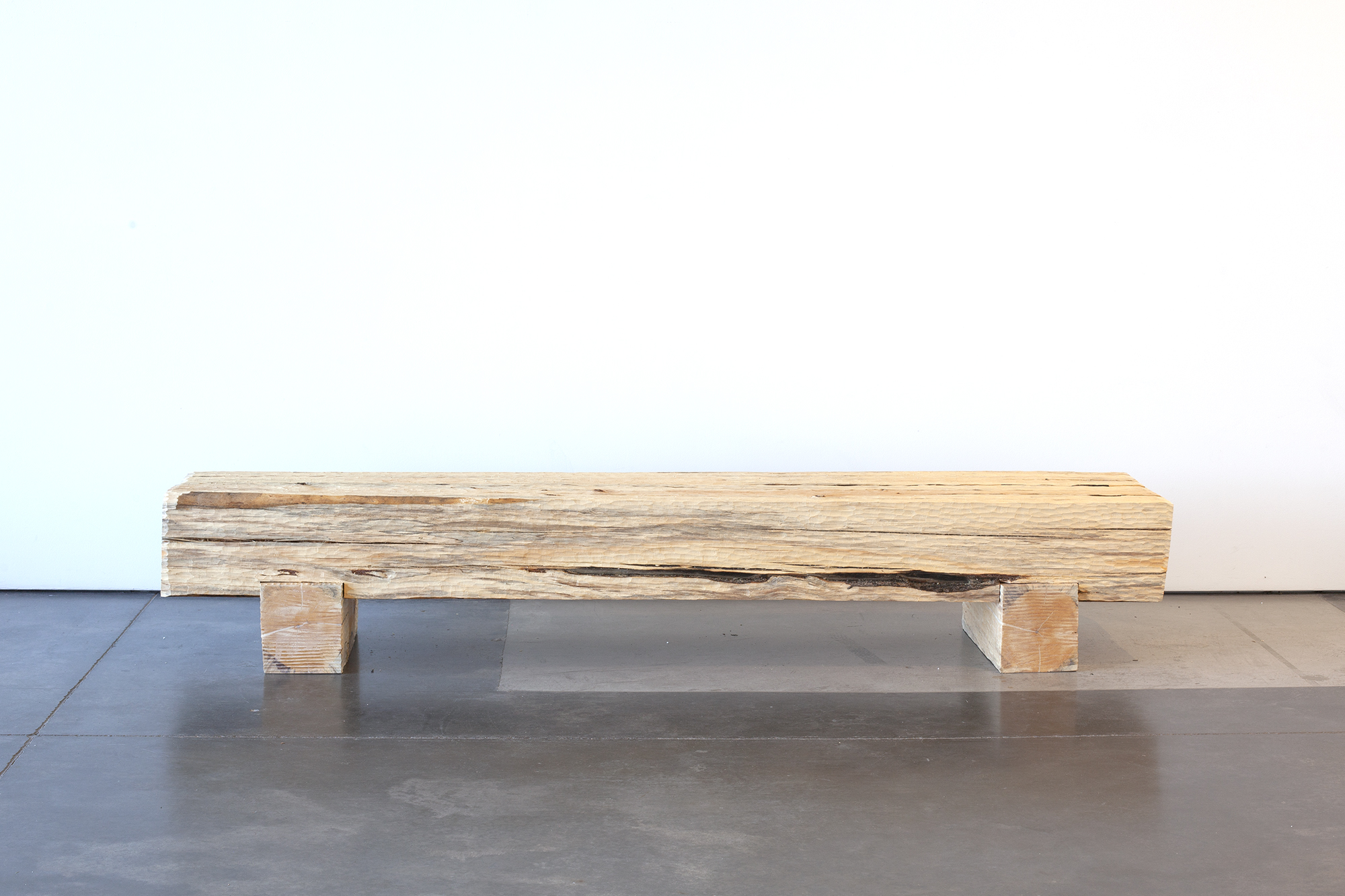 A long wooden bench made from a single weathered log with a dark burn scar running across its top surface, supported by two rectangular wooden blocks, sits on a polished concrete floor against a white wall.
