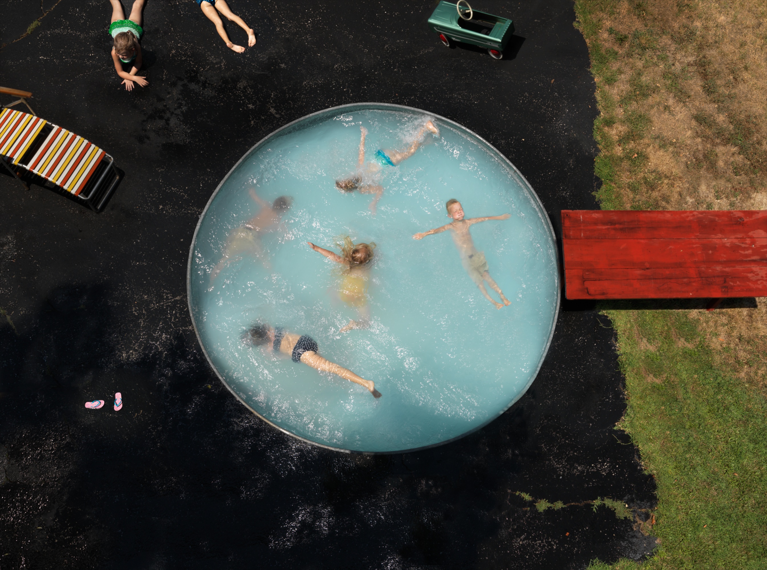 Aerial view of children swimming and playing in a large circular stock tank filled with turquoise water, surrounded by a dark asphalt surface with a red wooden bench, striped lounge chair, toy truck, and children standing nearby on grass and pavement.