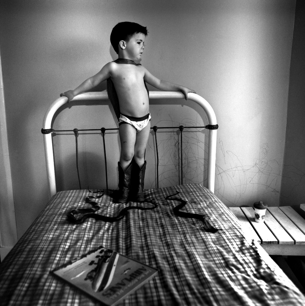 A young child stands on a bed wearing boots and underwear, gripping the metal headboard rails with arms outstretched. The black and white photograph captures the figure in profile against a plain wall, with a plaid bedspread, scattered drawings, and household items visible on the mattress surface.
