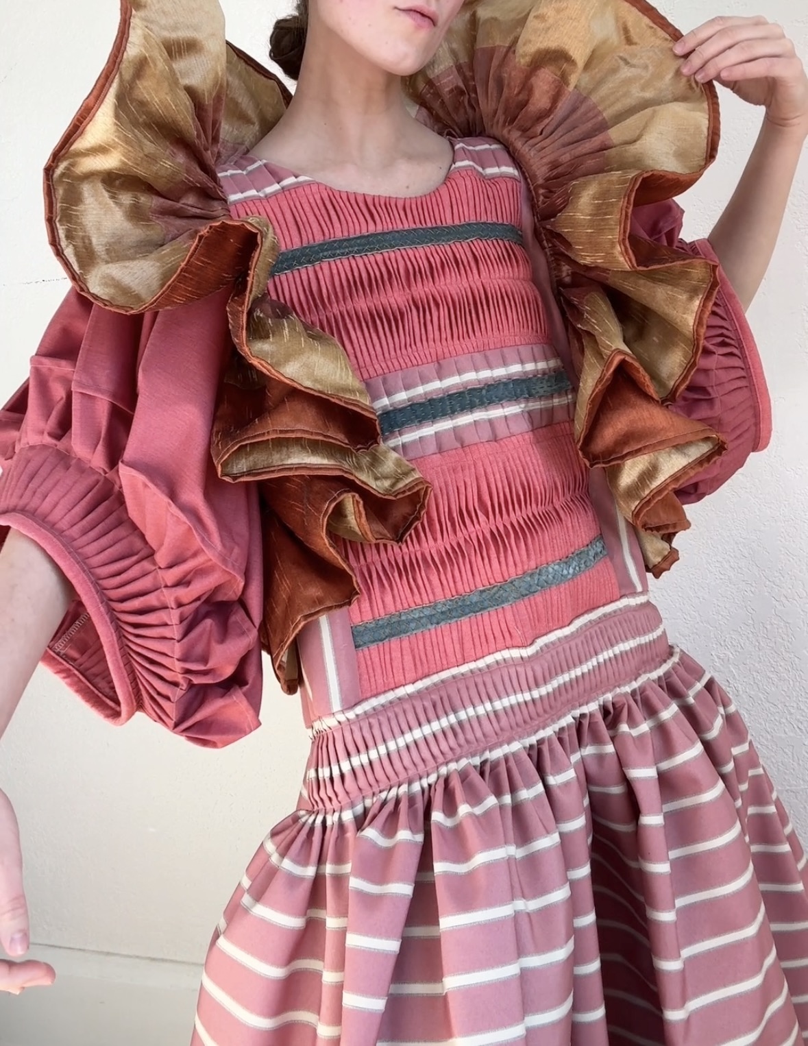 A figure wearing a voluminous pink dress with horizontal striped pleating and white banding stands with arms raised, displaying dramatic ruffled shoulders in contrasting gold and burgundy taffeta. The composition showcases intricate textile craftsmanship through hand-pieced pleats and embroidered details throughout the garment.