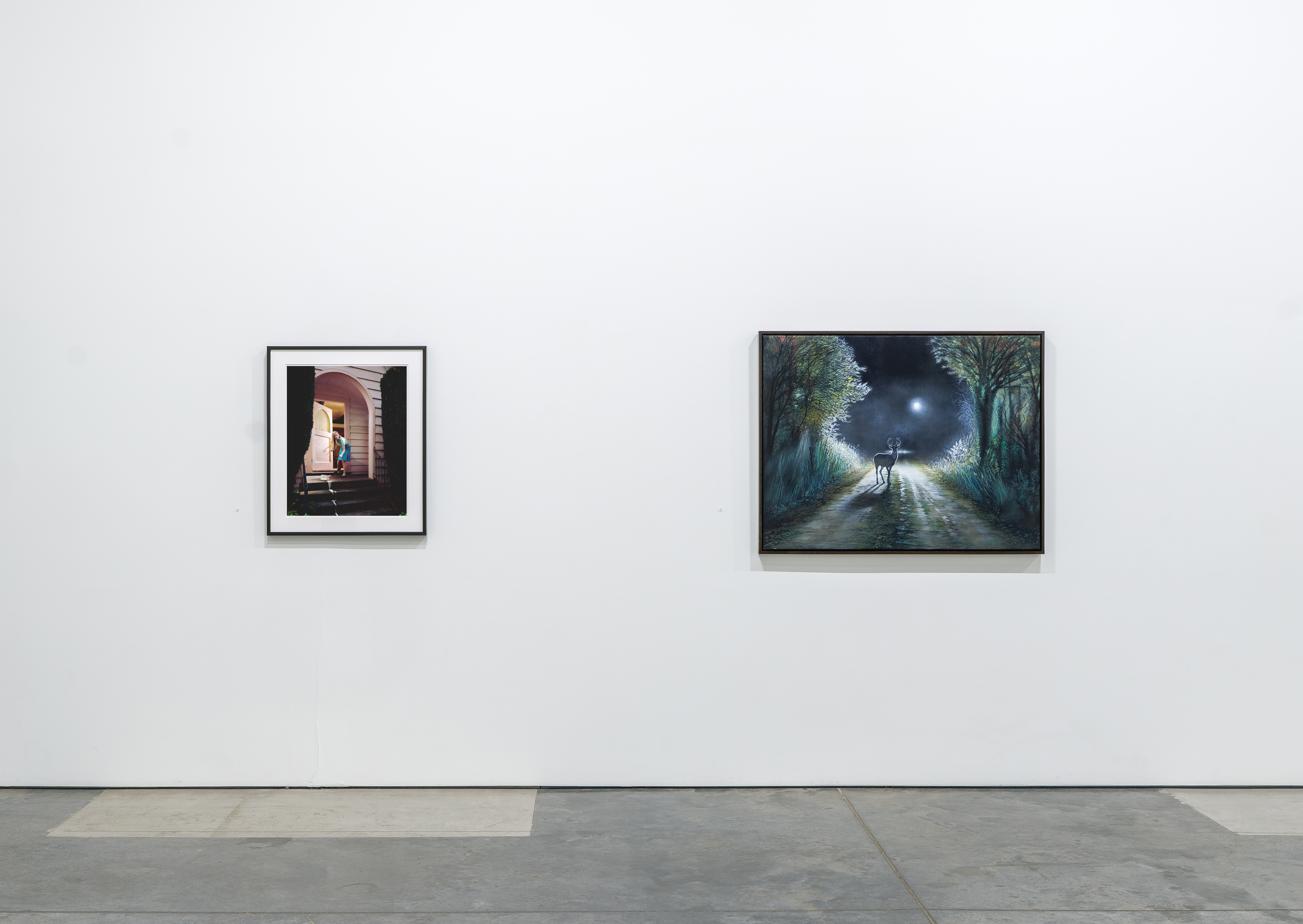 Gallery wall displaying two framed artworks: on the left, a smaller photograph with dark borders showing a figure in an illuminated archway; on the right, a larger landscape painting depicting a solitary figure standing on a moonlit rural path flanked by trees.
