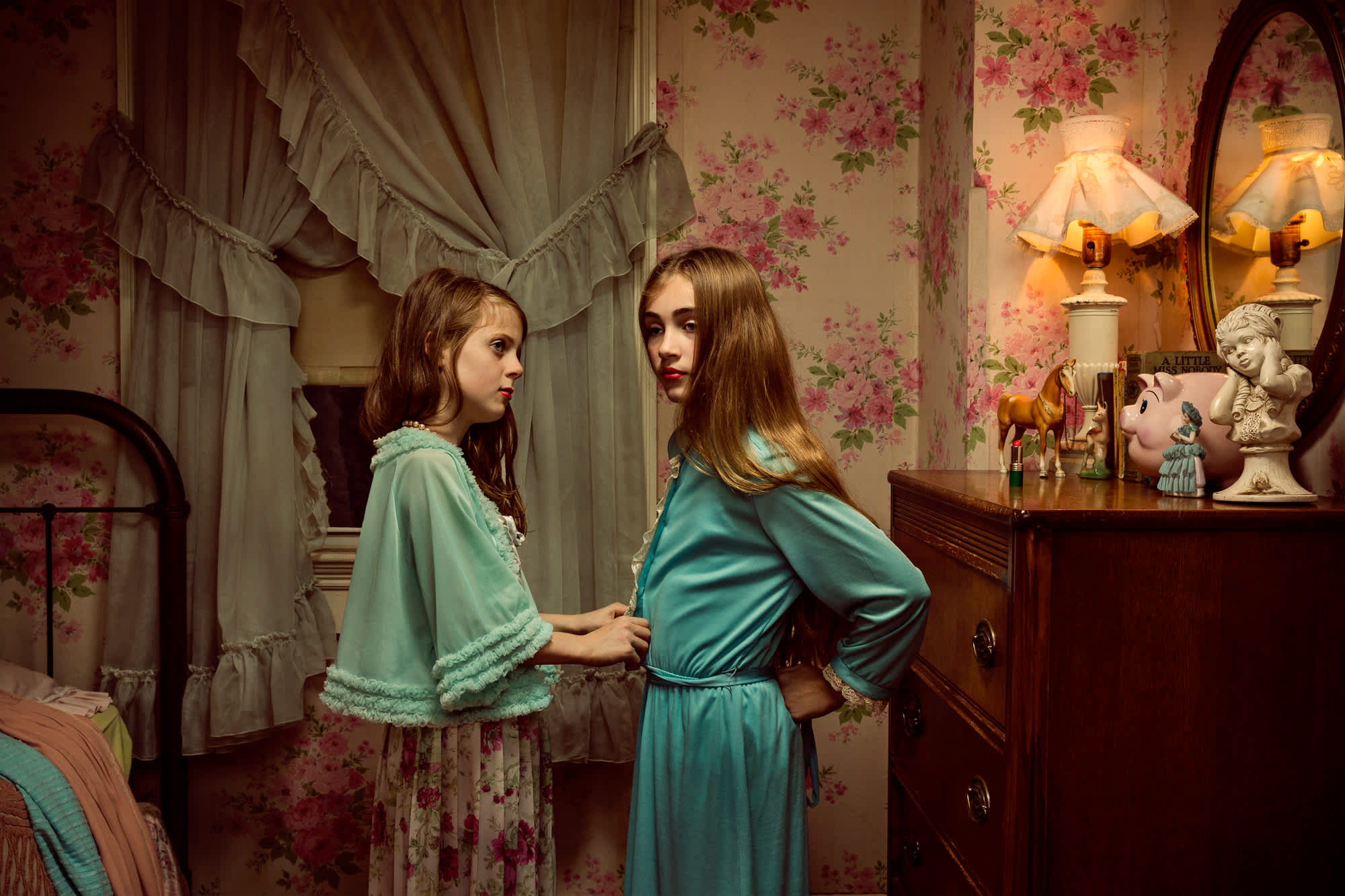 Two young girls in turquoise robes stand facing each other in a vintage bedroom decorated with rose-patterned wallpaper and sheer curtains. Warm lamplight illuminates classical decorative objects on a wooden dresser behind them, creating a nostalgic, staged tableau.