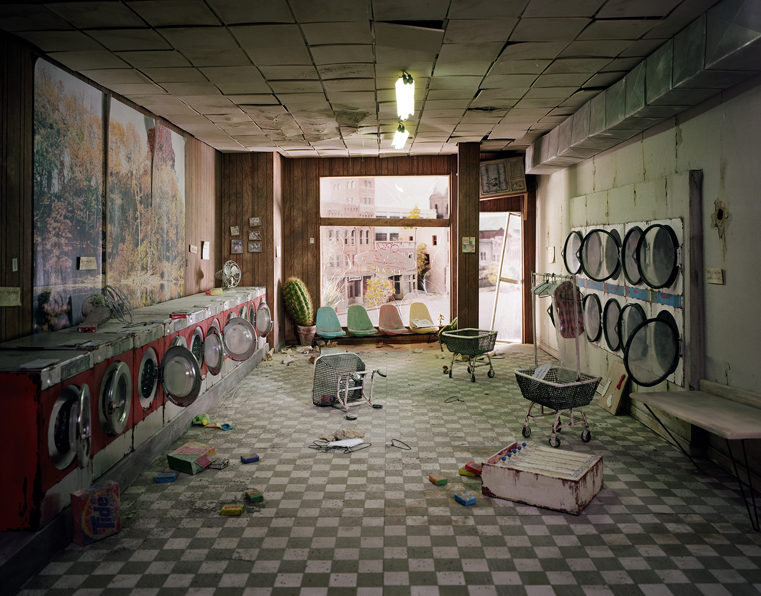 An abandoned laundromat interior with red and black washing machines lining the left wall and dryer units mounted on the right. The checkered floor is scattered with toys and debris, while colorful plastic chairs and laundry baskets sit near large windows overlooking an urban cityscape. Fluorescent ceiling lights illuminate the deteriorating space with peeling paint on pale green walls.