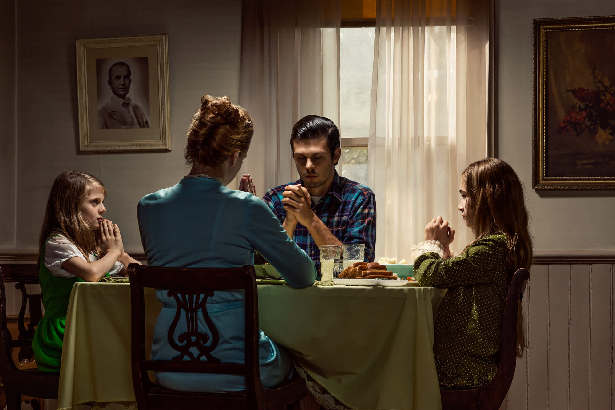 A family of four sits around a dining table in a formal interior room with framed portraits on pale walls and sheer curtains filtering natural light. The figures wear earth-toned and jewel-toned clothing; two younger figures sit on the left and right while an adult in a teal shirt faces away from the camera, and another adult in a plaid shirt sits between them. Glasses of water and food are visible on the table covered with a sage green cloth.