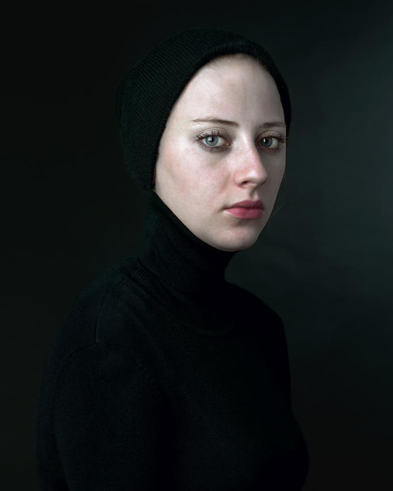 Portrait of a pale-skinned person wearing a black knit hood and turtleneck, photographed against a dark background in three-quarter view. The subject gazes downward with a contemplative expression, their face luminously lit against the shadowed composition.