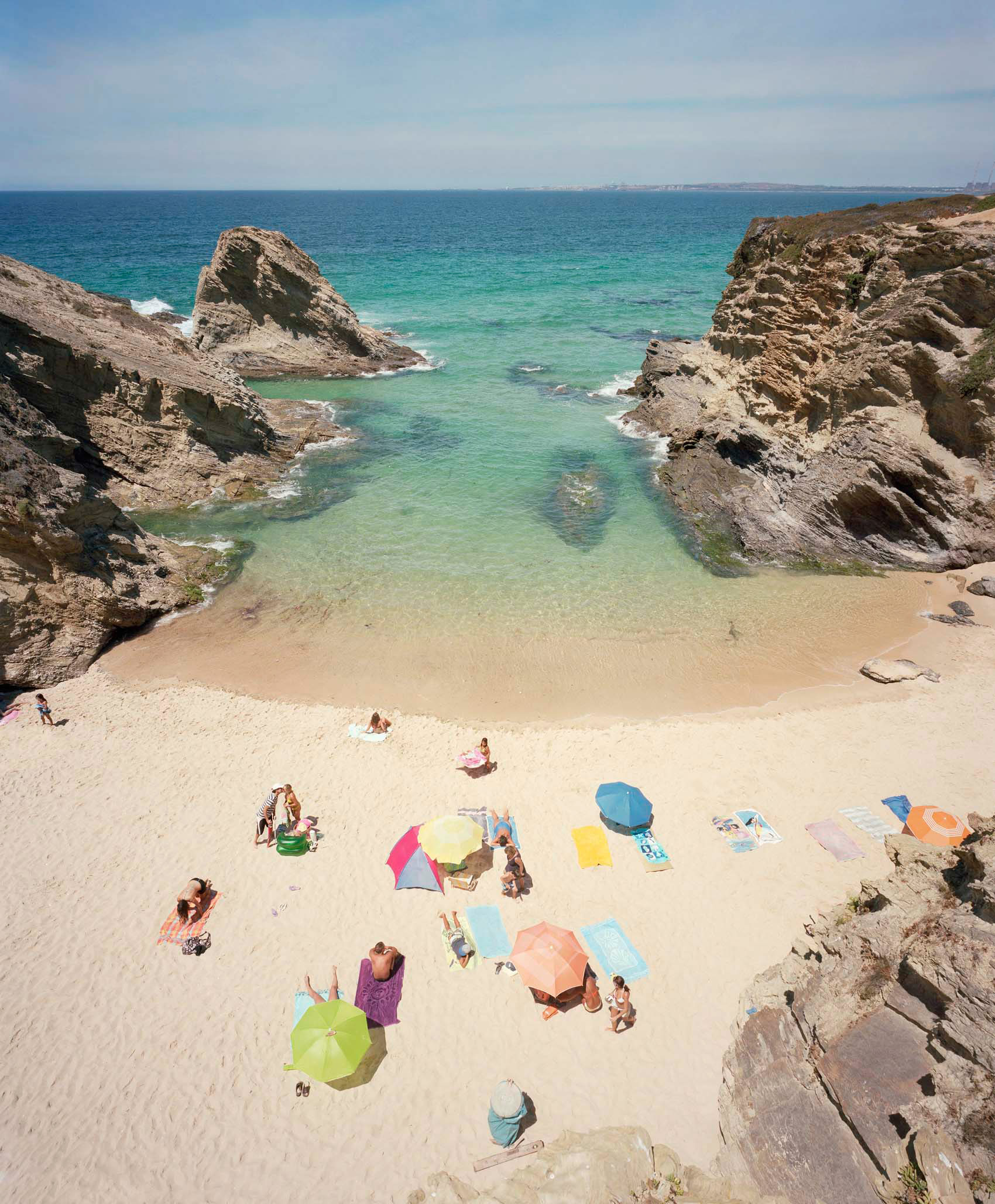 Aerial view of a secluded beach cove framed by dramatic rocky outcrops, with crystal-clear turquoise water and golden sand. Colorful beach umbrellas and sunbathers are scattered across the shore, creating vibrant pops of pink, yellow, blue, and green against the natural landscape.