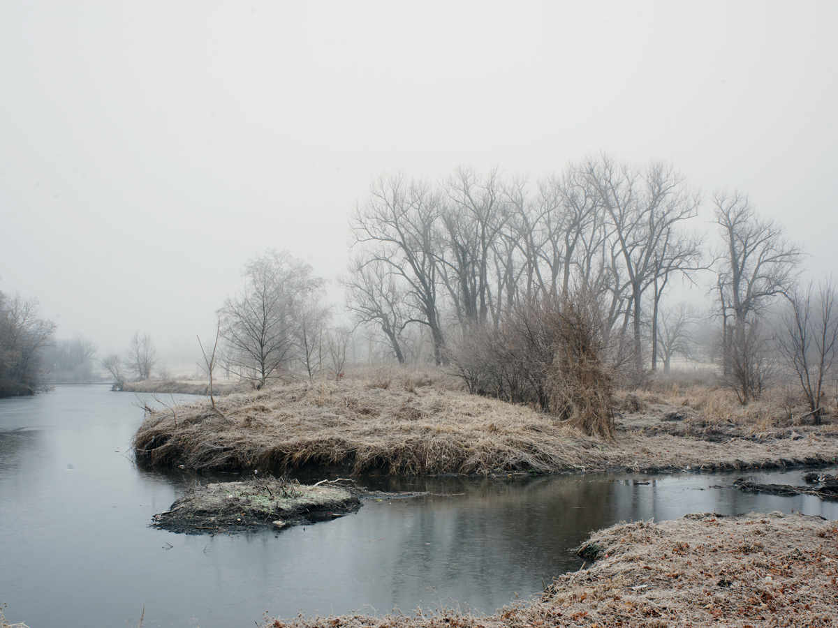 A misty riverside landscape with bare, leafless trees receding into fog. The composition features a shallow creek or waterway in the foreground with sandy banks and rocky outcrops, bordered by dormant vegetation and a dense stand of skeletal trees along the far bank. The overall palette is muted in grays, browns, and pale tones, creating a somber, winter atmosphere.