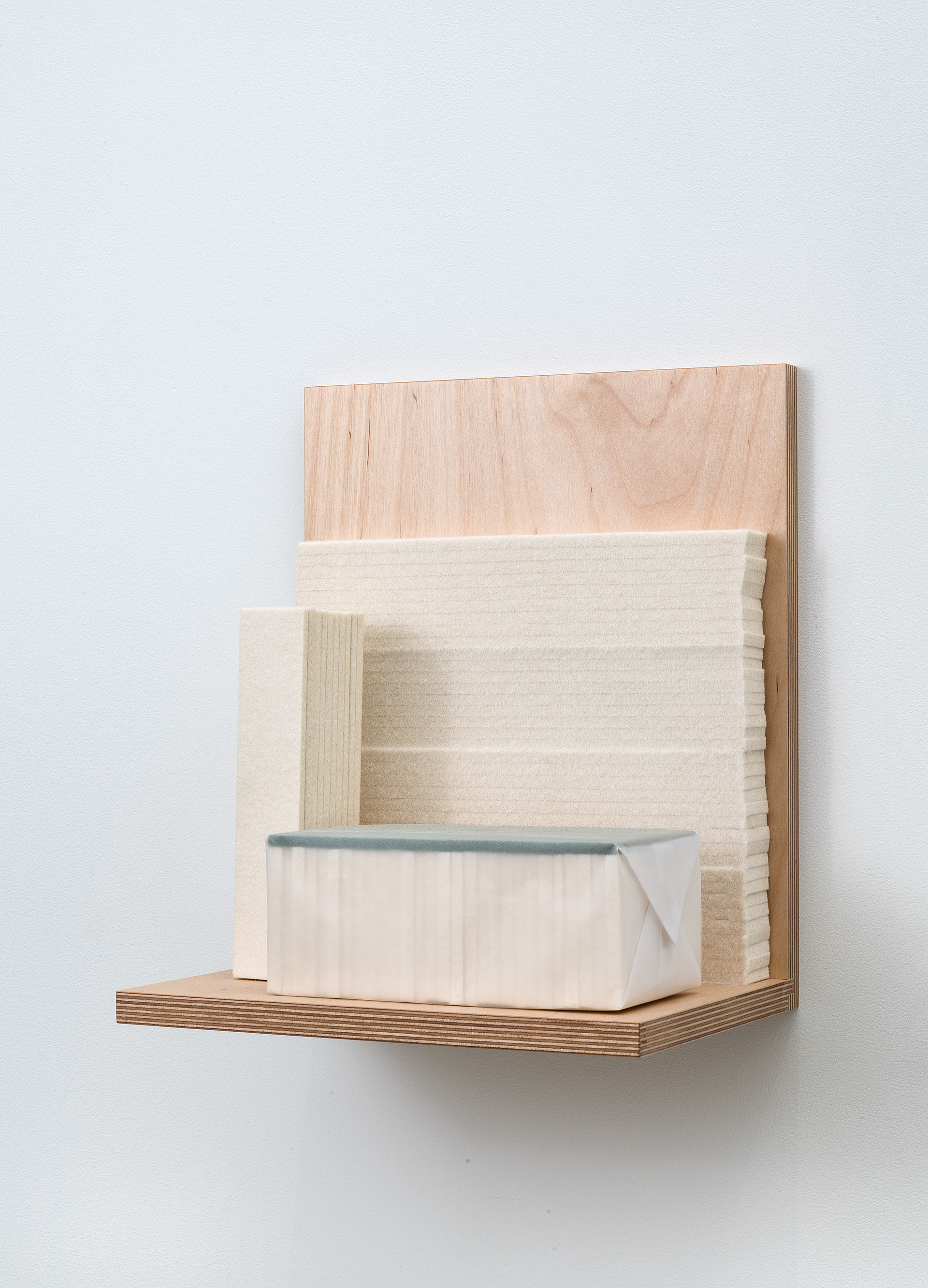 A wooden wall-mounted shelf unit constructed from light plywood and natural wood veneer, featuring geometric compartments and a cream-colored fabric-wrapped box with a blue-gray felt edge resting on the lower shelf.