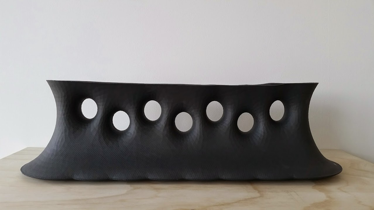 Black porcelain vessel with a undulating, curved form featuring seven evenly-spaced circular apertures across its front face, displayed against a white wall on a light wooden surface.