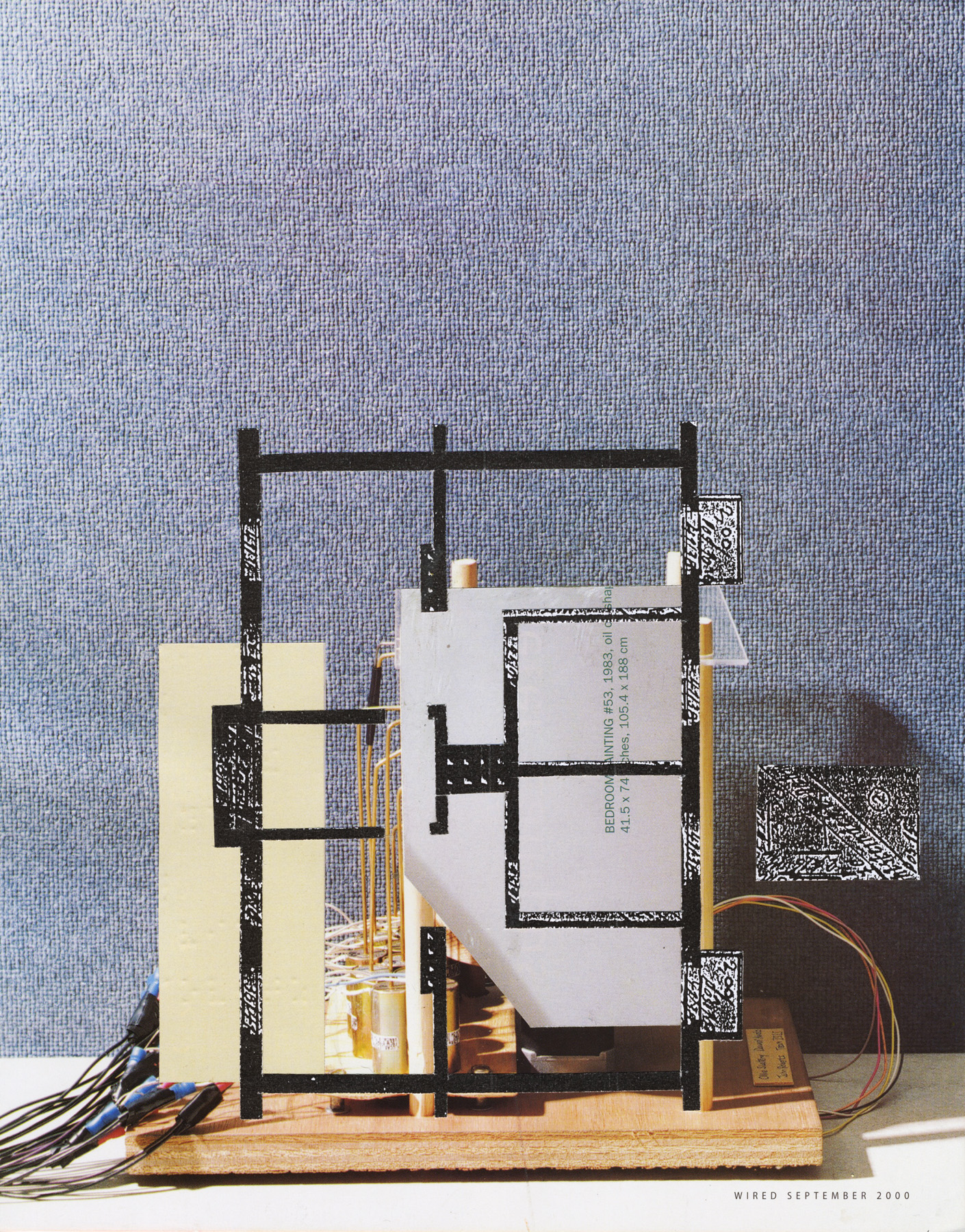 A three-dimensional collage construction mounted on a wooden base features black linear frameworks resembling architectural models, cream-colored paper planes, white rectangular forms with printed text, and wired electronic components in orange and red. Against a textured blue-gray backdrop, the assembled elements create a layered, abstract composition blending architectural, industrial, and mixed-media aesthetics.