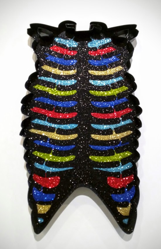 A skeletal ribcage rendered in black with symmetrical horizontal bands of glitter in vibrant colors—cyan, red, blue, gold, and chartreuse—suspended in glossy resin. The form is frontal and vertical with pointed ends at top and bottom, mounted against a white wall.