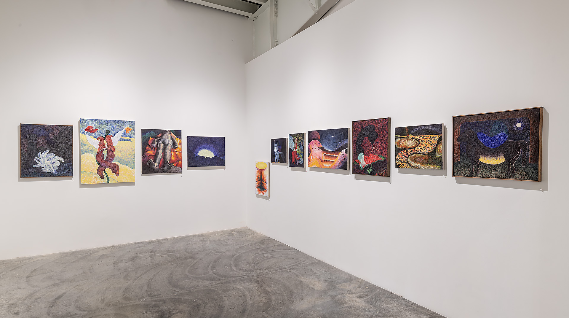 Gallery wall displaying a linear arrangement of richly textured paintings and a sculptural form on a concrete floor. The works feature vibrant reds, blues, golds, and earth tones with figurative and abstract imagery including celestial motifs, human forms, and organic shapes. A small vertical sculptural piece with warm tones stands centrally among the framed works, creating a rhythmic composition across the white wall.