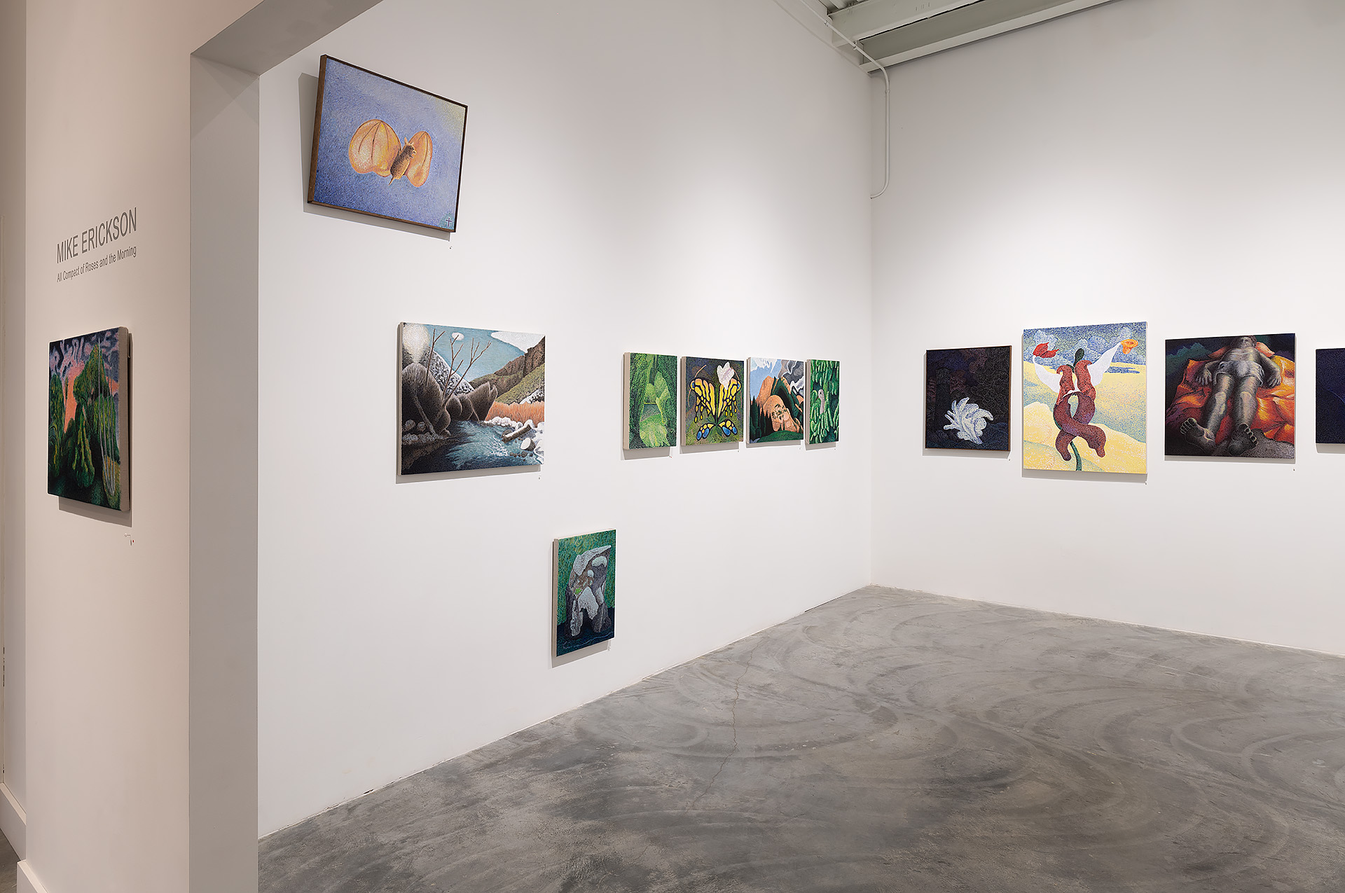 Gallery installation view featuring a collection of paintings arranged across white walls. Works include nature-inspired scenes with animals and landscapes rendered in rich colors—vibrant greens, blues, and earth tones—displayed at varying heights. A screen mounted above shows an image of peeled oranges on a blue background. Polished concrete flooring and minimalist gallery architecture frame the exhibition.