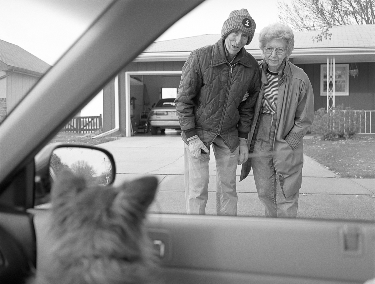Black and white photograph taken from inside a car, framing two people standing in a driveway in front of a residential house. A man wearing a quilted jacket and knit hat stands beside an older woman in glasses and a striped shirt. Both smile toward the camera as they stand on the concrete driveway, with a garage and bare tree branches visible in the background.