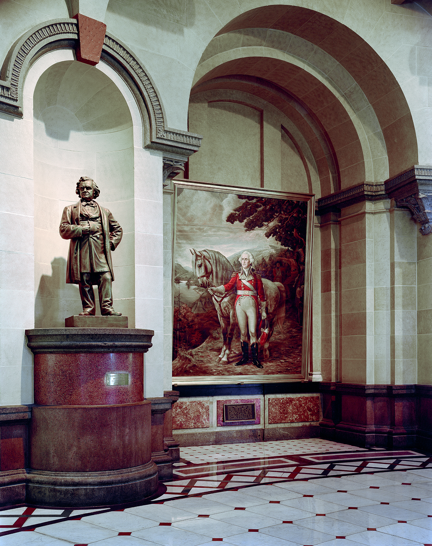 A neoclassical corridor with arched ceilings features a bronze statue on a cylindrical red marble pedestal on the left, facing a large framed historical painting depicting a uniformed figure on horseback. The geometric floor in cream and deep red tones, ornamental architectural details in muted greens and browns, and formal gallery lighting create a symmetrical, dignified interior space.