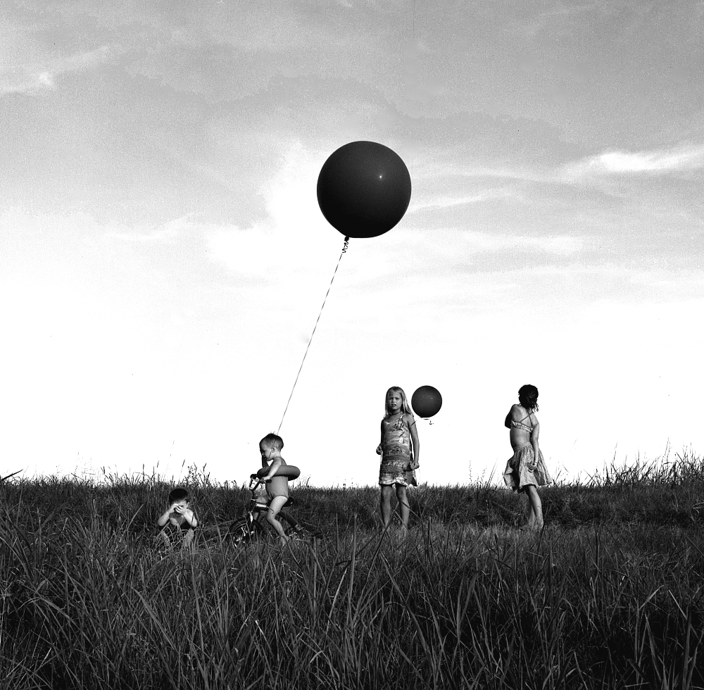 Black and white photograph of four children standing in a grassy field beneath a cloudy sky, with a large black balloon tethered to a string held by the child on the left, floating high above them.