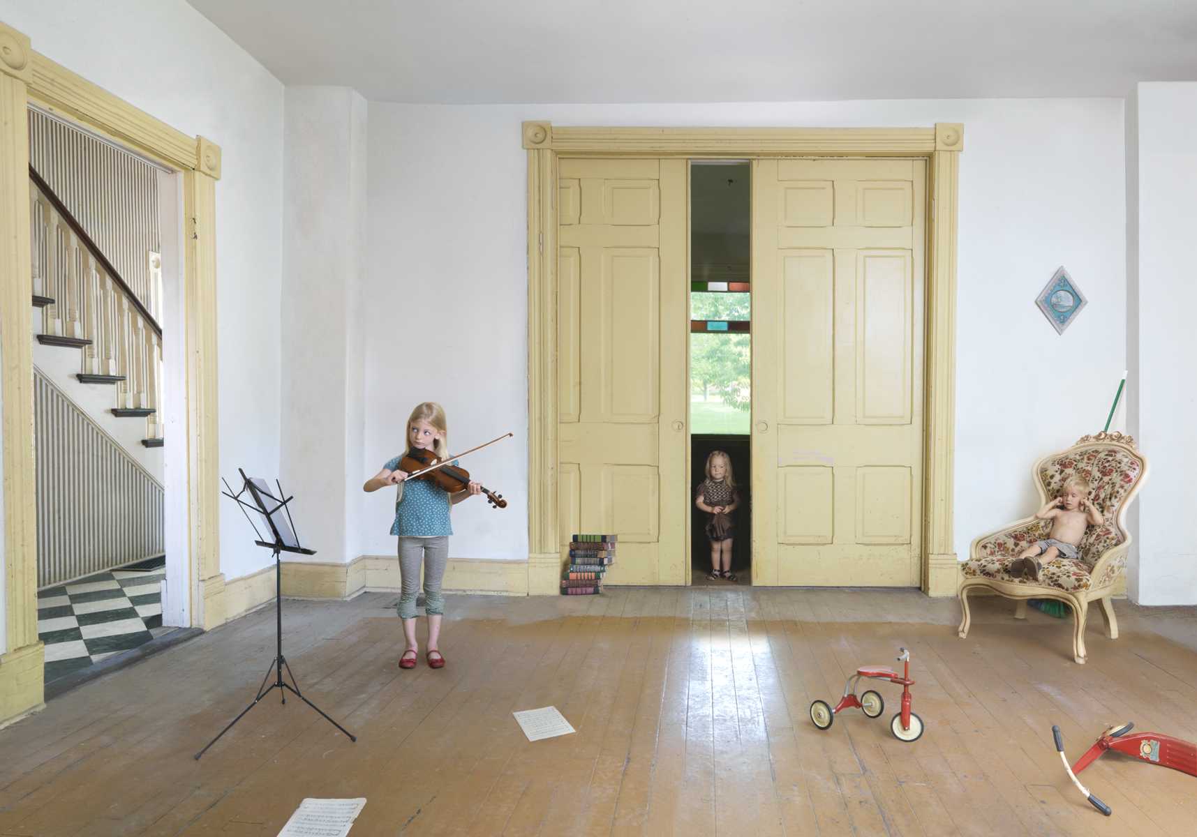 A young girl plays violin in a spacious, formally decorated room with pale yellow-trimmed doors and white walls. The composition includes a music stand, a red tricycle, an ornate upholstered chair, stacked books, and another child visible through the open doorway, arranged in a carefully composed domestic interior scene.