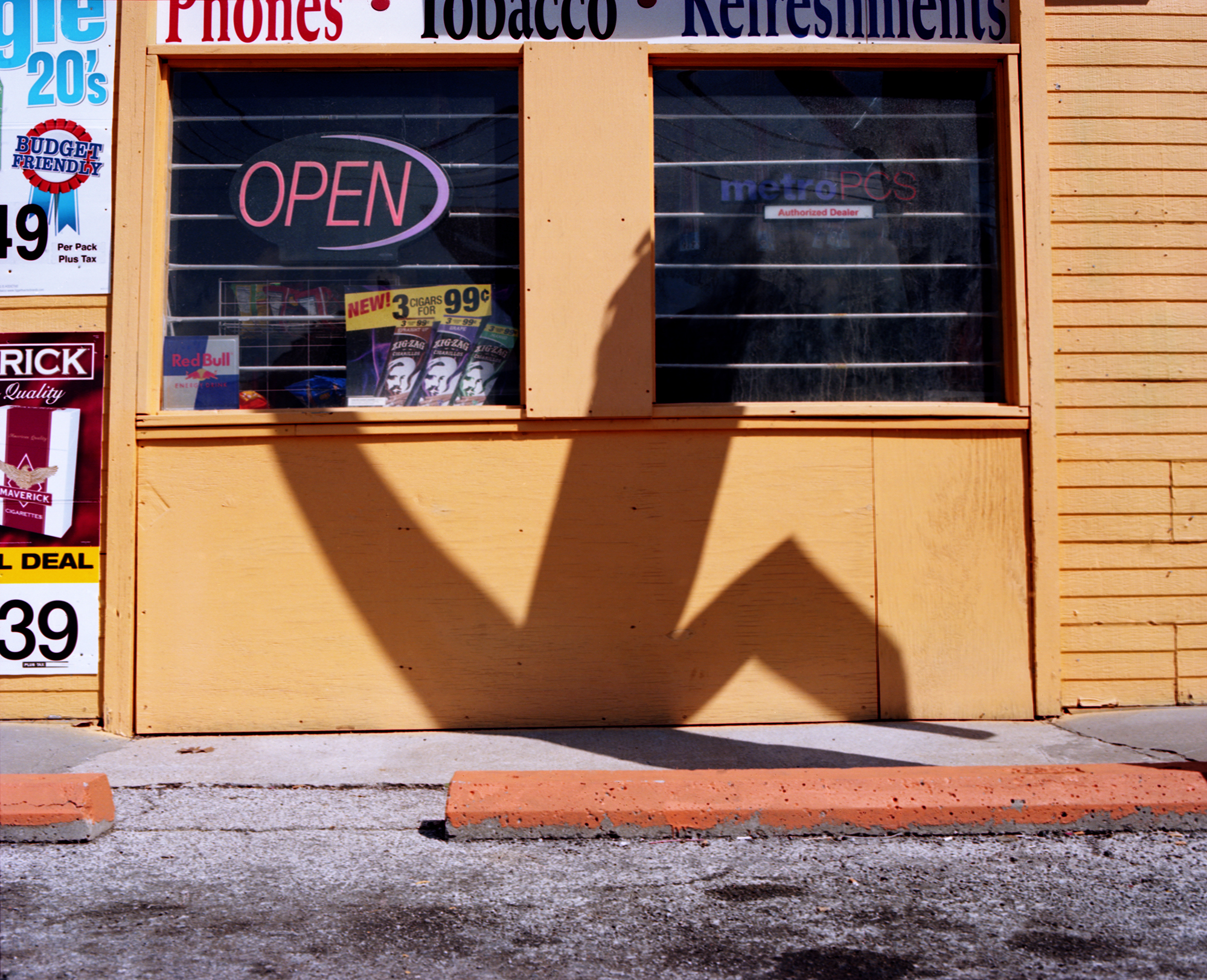 A storefront with tan wooden siding and two large windows displays an "OPEN" neon sign in pink. The harsh midday sun casts a distinct shadow of a hand with fingers spread across the yellow facade and lower window area, creating a striking contrast against the warm-toned building.