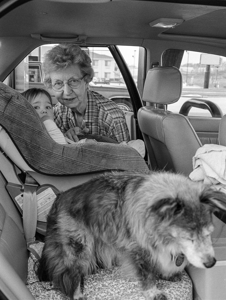 Black and white photograph taken from inside a car showing an elderly woman with glasses holding a young child in the back seat, with a long-haired dog in the foreground. Street and buildings are visible through the car windows.