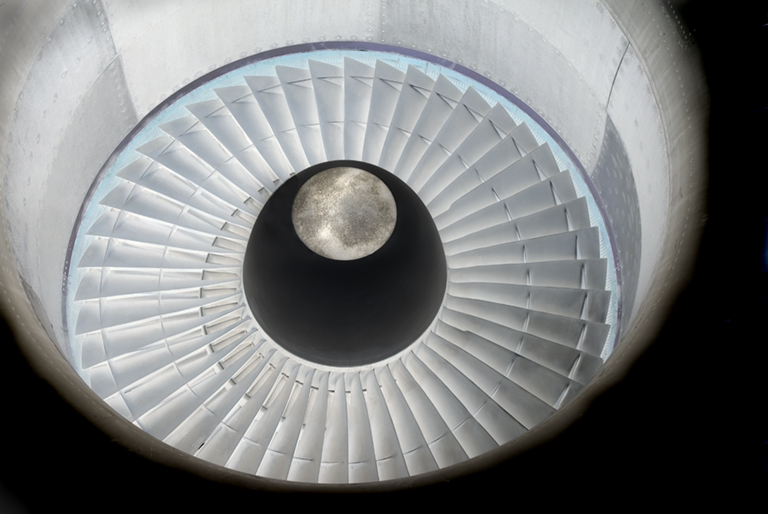 Overhead view of a circular architectural interior featuring radiating white panels that create a spiral pattern, with a dark spherical void at the center topped by a luminous circular form suggesting moonlight or a celestial body.