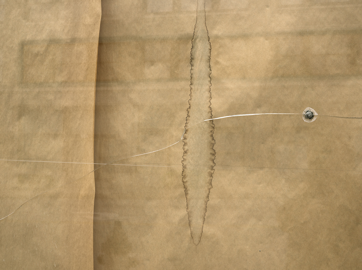 Folded tan paper with a vertical torn seam down the center, accompanied by two thin translucent lines extending across the composition, each terminating at a small spherical form on the right side.