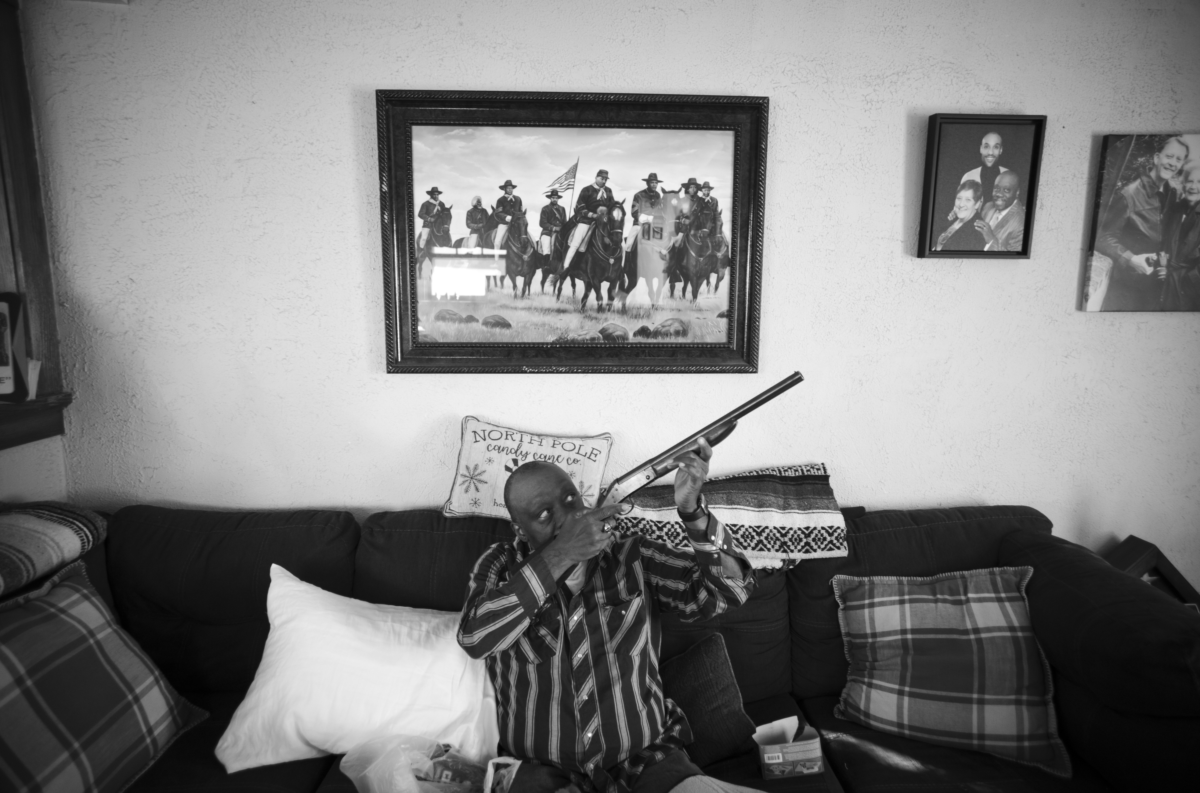 Black and white photograph of a person seated on a dark couch holding a rifle, positioned beneath framed photographs and artwork on a white wall. The composition captures an intimate domestic interior with patterned pillows and blankets, while the subject's pose creates a stark juxtaposition between the domestic setting and the weapon.
