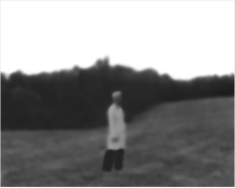 A solitary figure in a white shirt and dark pants stands in a vast, empty field, facing away from the camera toward a dark treeline on the horizon. The black and white photograph has a soft, slightly out-of-focus quality that emphasizes the isolation of the lone person in the expansive landscape.