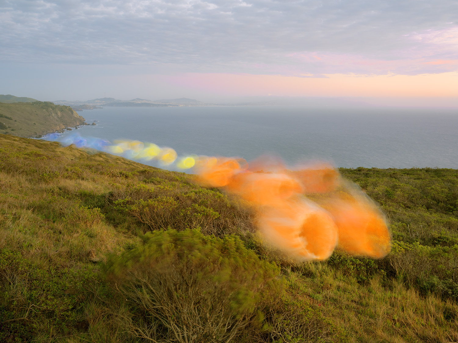 Coastal landscape with rolling green hills descending toward a calm sea, featuring a trail of colorful circular forms—blue, yellow, and orange—overlaid across the terrain in a diagonal composition, with distant headlands visible under a soft, overcast sky.