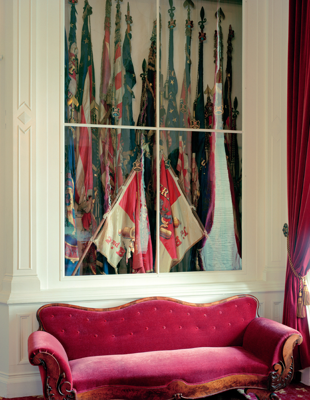 A framed photographic print depicting a collection of ornate flags and banners in jewel tones—greens, reds, and blues—displayed vertically. Below the print sits a luxurious magenta velvet settee with curved arms and decorative nail head detailing, positioned against a cream-colored wall with red velvet curtains visible to the right.