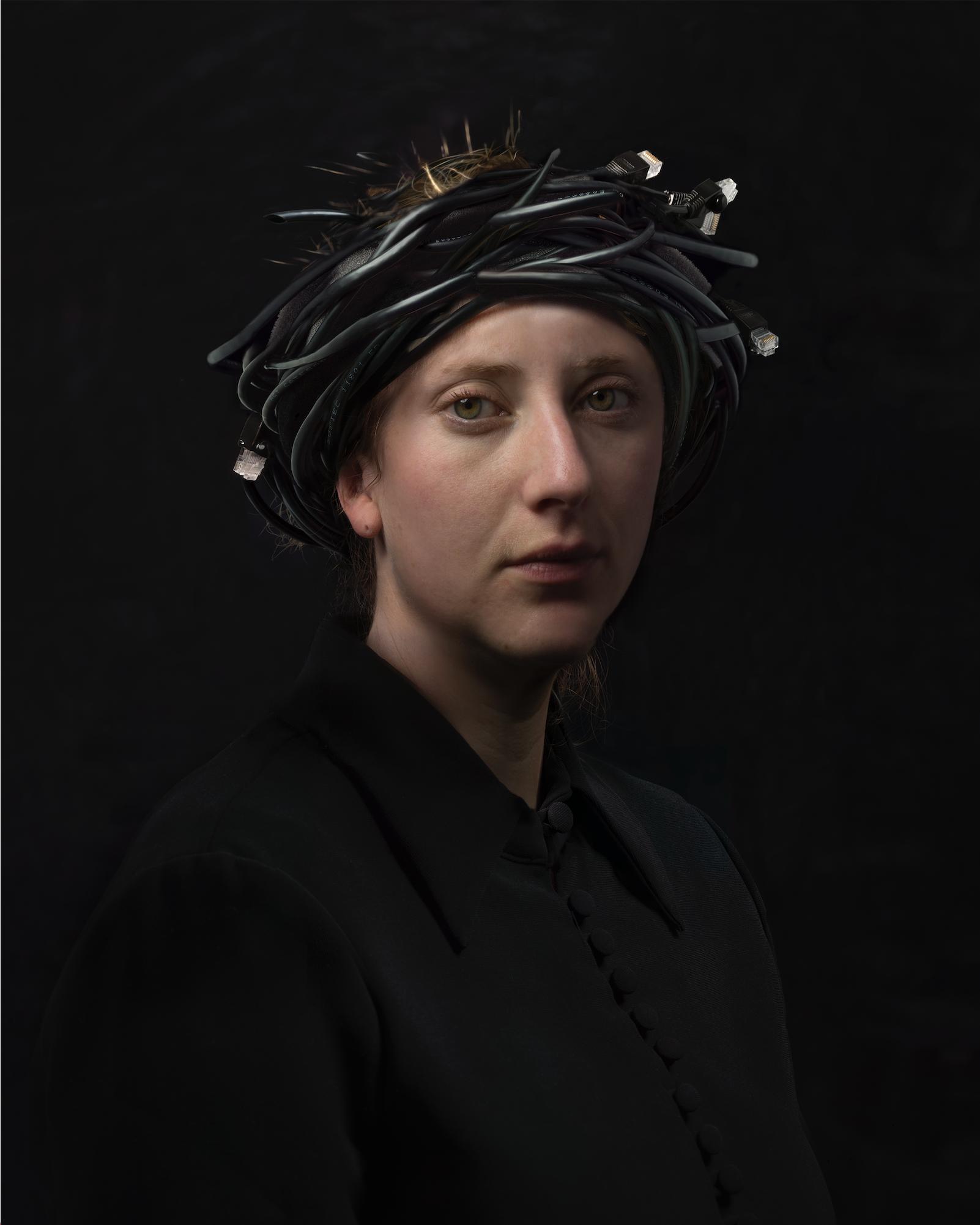 Portrait of a figure with pale complexion against a dark background, wearing black clothing and an elaborate headdress constructed from black cords, straw, and metallic elements. The composition is a three-quarter view with dramatic lighting that emphasizes the subject's contemplative expression and the intricate textural details of the headpiece.