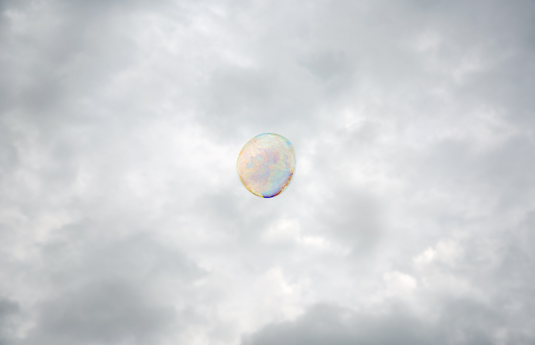 A translucent iridescent sphere floats against a soft, overcast sky filled with white and gray clouds. The bubble's delicate surface displays subtle shifts of pastel color—pale blues, pinks, and yellows—characteristic of light refracting through a thin film.