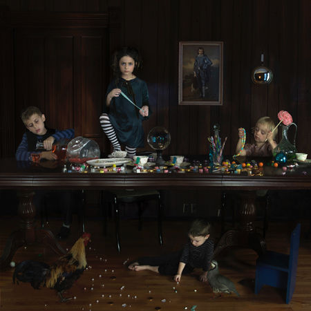 Four children gather around a dark wooden table laden with art supplies and scattered candies, while one child reaches down to collect sweets from the floor below. The scene is set in a dimly lit interior with dark walls, a framed portrait visible in the background, and warm overhead lighting that creates a theatrical, staged quality to the composition.